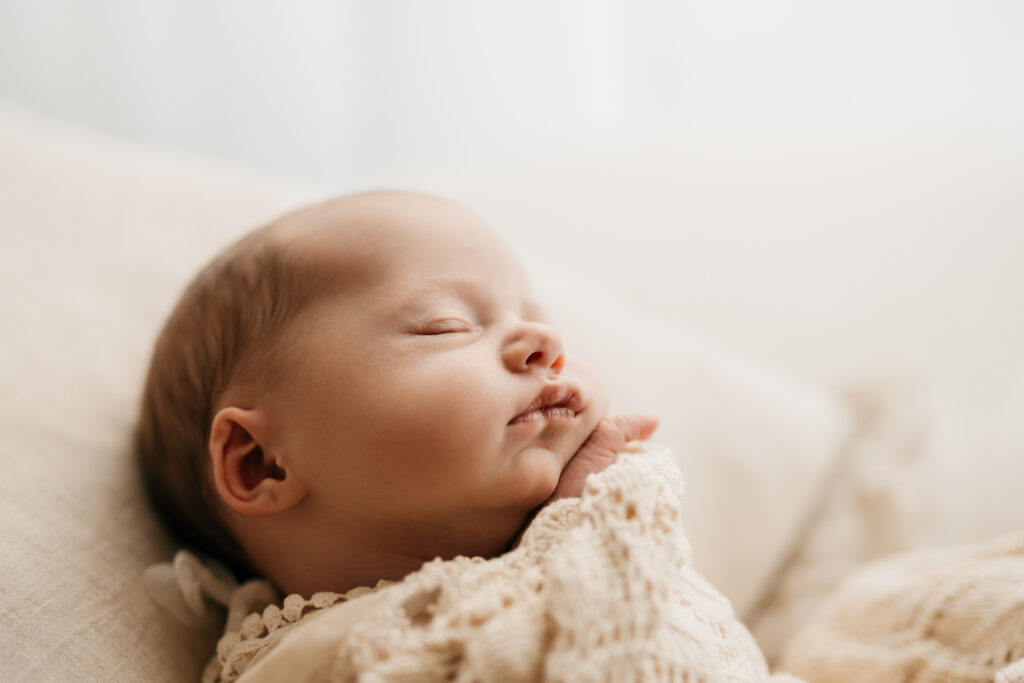 Photo of a newborn baby asleep with her hand under her chin, wrapped in a blanket demonstrating natural newborn photography posing. 
